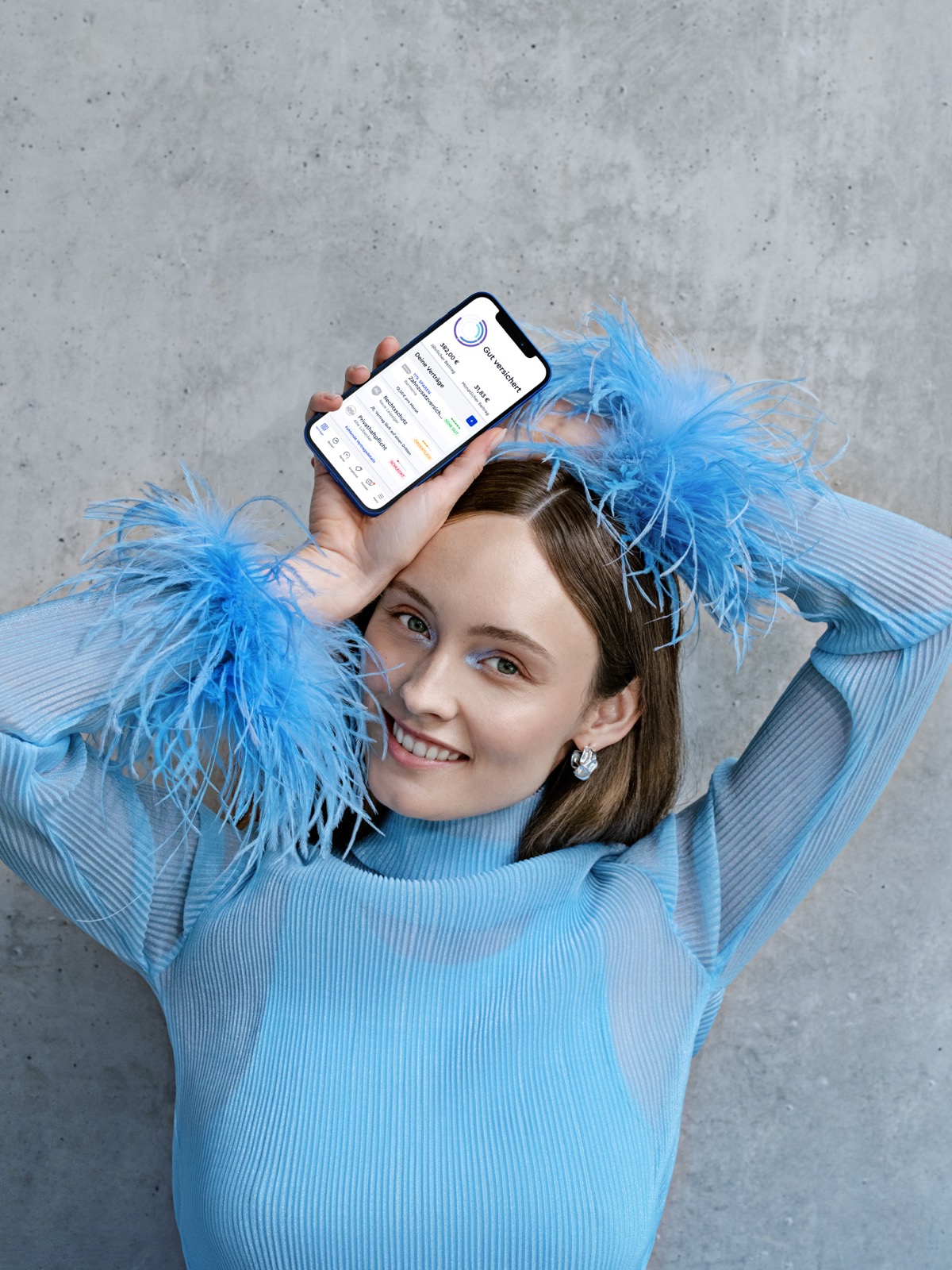 CLARK product photography — smiling model in a blue feathered top holding the phone above her forehead against concrete